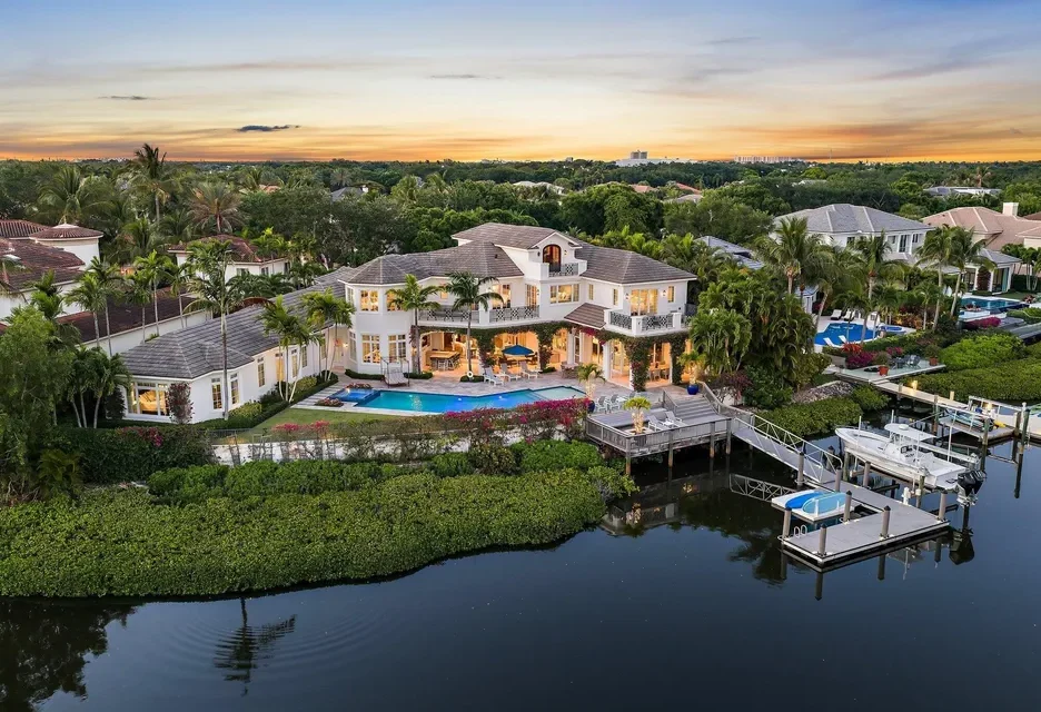 Aerial view of a waterfront home in Jupiter, FL, surrounded by lush greenery and palm trees at sunset.